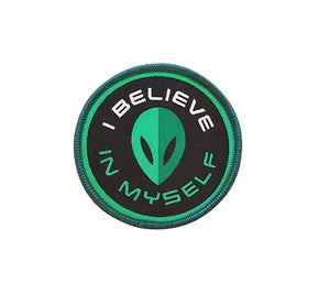 A circular iron on patch that is black, white, and shades of green. The patch has an alien face in the centre with curved text around it that reads "I believe in myself".