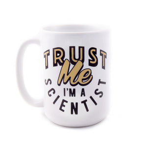 A white ceramic mug with curved black and gold script that reads "Trust Me, I'm A Scientist"